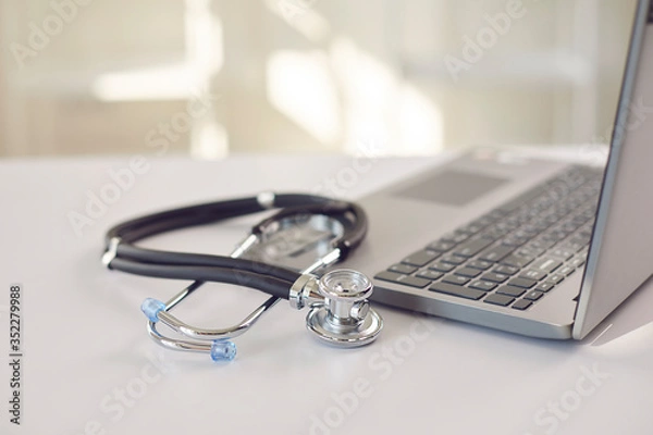 Fototapeta Doctor online. A stethoscope is on a white table with a laptop in blur at the hospital office.