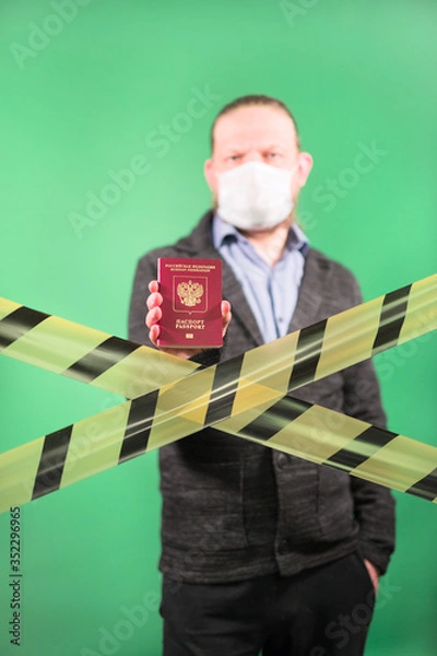 Fototapeta A man in a protective medical mask on a green background holds a passport in his hand. Yellow forbidden tape in front of a standing person. Closing borders due to the epidemic of coronavirus.