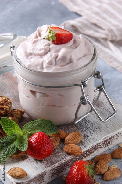 Fototapeta Fermented milk product. Healthy strawberry yogurt in a transparent jar on a white wooden board with fresh berries, mint, nuts and cookies on a light grey background. Background image, copy space