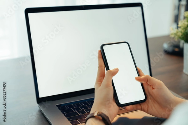 Fototapeta Woman hand holding smartphone with blank copy space screen for your advertisement. laptop on the table.