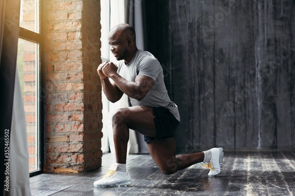 Obraz Muscular african american trainer in tight t-shirt doing an exercise lunging forward while holding hands at chest