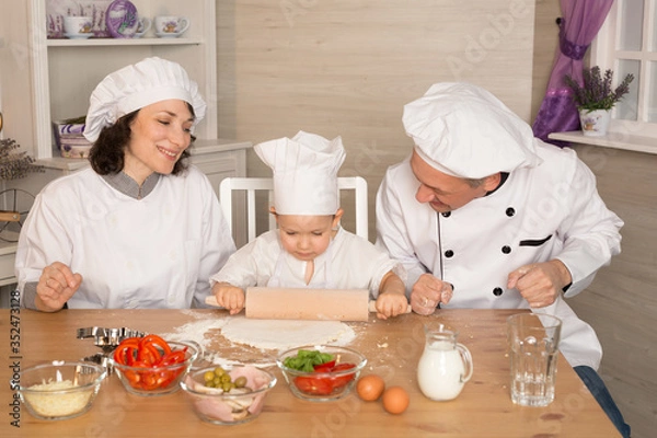 Fototapeta Mom, dad and little son cook together in quarantine. The family laughs and cooks pizza in cookery suits. A small child rolls out the dough with a rolling pin.
