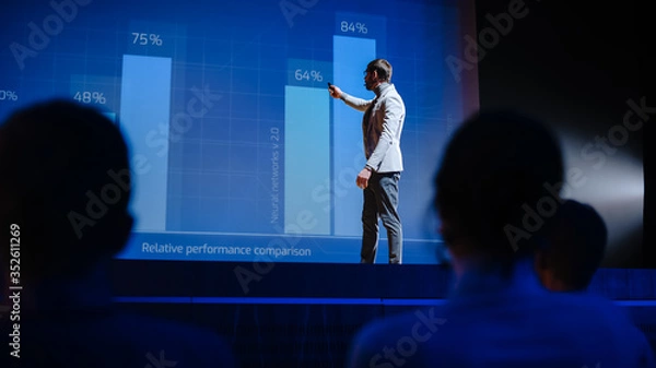 Fototapeta On Stage Visionary Male Speaker Presents New Technology, Uses Remote Control for Presentation, Showing Infographics, Statistics Animation on Screen. Live Event / Start-up Business Conference