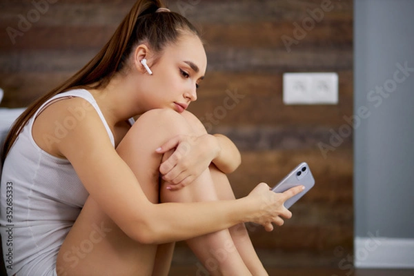 Obraz young and beautiful caucasian lady has rest at home, she sits on the floor next to bed and listen to music in headphones, using smartphone