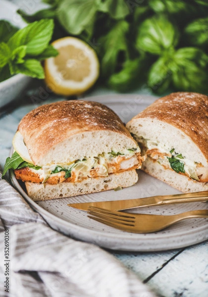 Fototapeta Breakfast table with fresh fried fish sandwich with tartare sauce, lemon and arugula cut in halves, fresh greens on plate over rustic wooden table. Healthy easy breakfast ideas