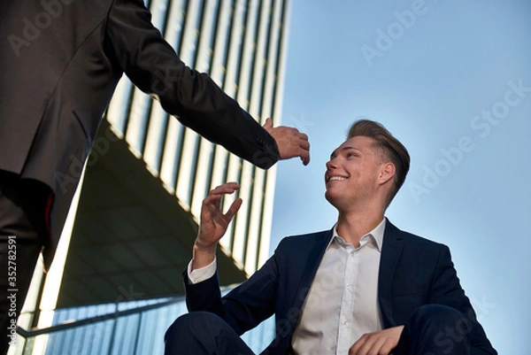 Obraz A young and an old businessman sit on the stairs between two high-rise buildings and celebrate your success, and shake hands,