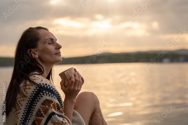 Obraz A charming dark haired young woman is sitting on the pier in a warm knitted poncho and sipping tea from a ceramic mug with her eyes closed