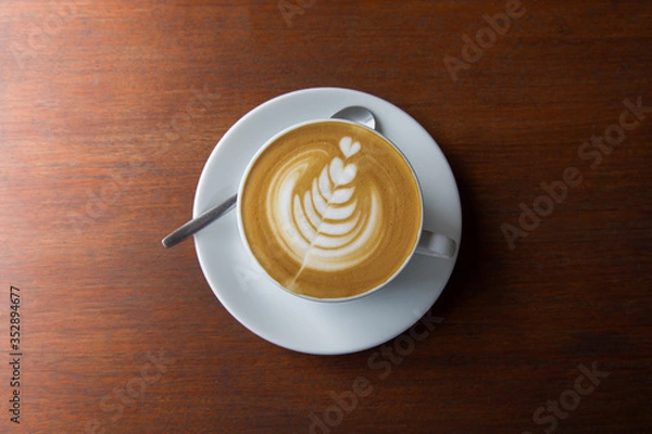 Fototapeta Top-view shot of single cup of latte coffee with art cream foam in white ceramic cup on wooden vintage table with sun lighting and shade in the morning after breakfast for relaxing and starting a day.