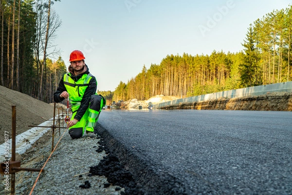 Fototapeta Surveyor engineer with equipment (theodolite or total positioning station) on the construction site of the road or building with construction machinery background