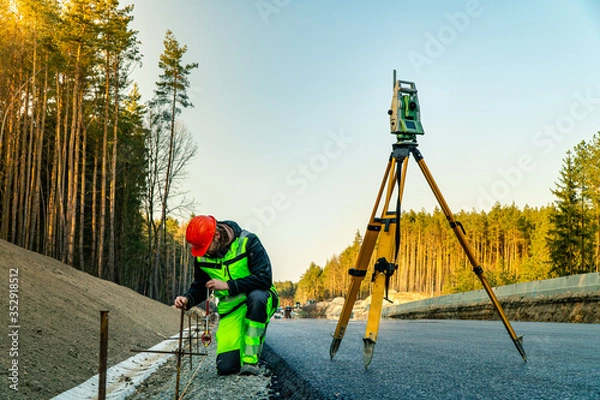 Fototapeta Surveyor engineer with equipment (theodolite or total positioning station) on the construction site of the road or building with construction machinery background