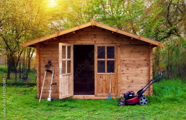 Fototapeta Hovel after work in evening, golden hour. Garden shed (front view) with hoe, string trimmer,  rake and grass-cutter. Gardening tools shed. Garden house on lawn in the sunset. Wooden tool-shed.
