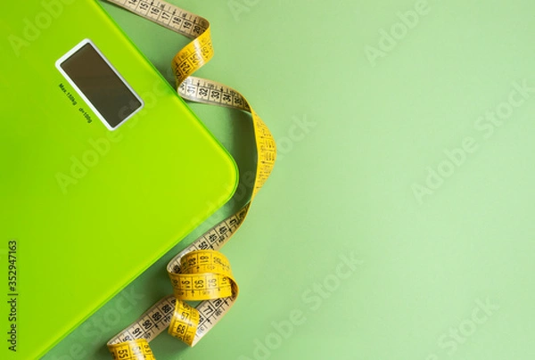 Fototapeta Green scales and yellow measuring tape on a green background. The concept of normal weight and health control. The horizontal position, lay flat, copy space