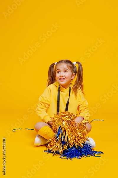 Fototapeta cute cheerleader girl in a yellow tank top and blue shorts sits with pompons in her hands