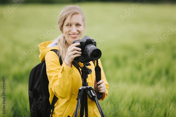 Obraz Portrait of attractive woman in eyeglasses and yellow jacket standing among wheat field with tripod and digital camera. Smiling female photographer enjoying spring weather outdoors.