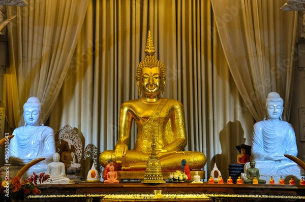 Fototapeta Golden Buddha statue with two white Buddhist statues on both sides on the beige curtain background. Buddhism concept.