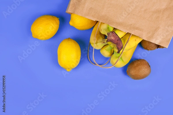 Fototapeta Bananas, kiwi and lemons in a paper eco friendly bag on a purple background. Eco shopping concept.
