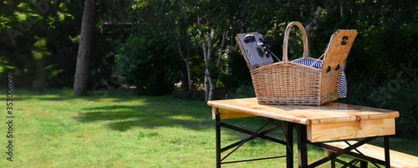 Fototapeta Open picnic basket on wooden table in green park, space for text. Banner design