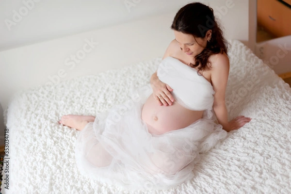 Obraz Active pregnant woman sitting on bed and touching her belly at home. A pregnant smiling girl is resting on a white bed. The last months of pregnancy.