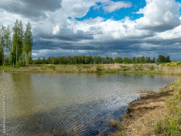 Fototapeta a fish pond, in the water of which there are colorful cumulus clouds and trees, there are recently dug gravel piles on the shore of the pond, the first spring greenery in nature