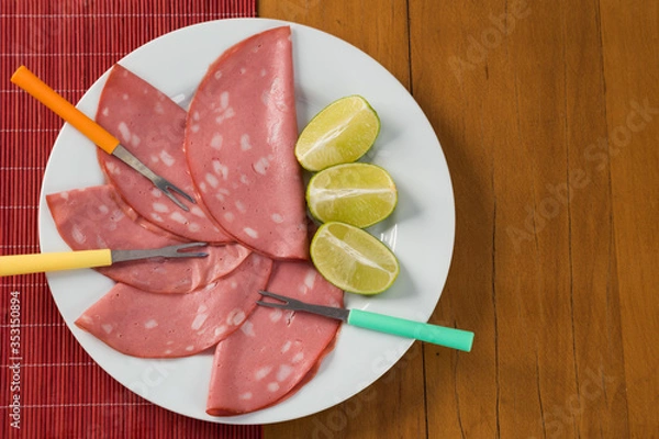 Fototapeta slices of mortadella served with lemon on a white plate, on a wooden background. top view