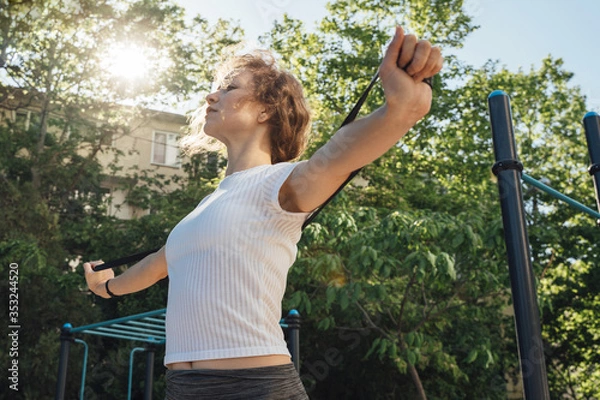 Fototapeta A woman in grey leggings and a white top looks in front of her and performs an exercise in her arms together to frame the waist with a sunny glare from behind