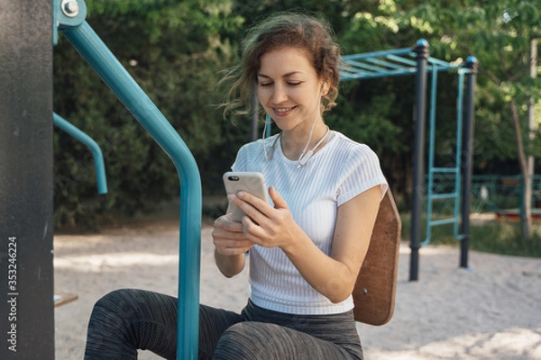 Fototapeta Girl brunette with blue eyes in a white top looks into the mobile phone and listens to music middle plan