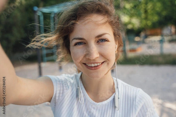 Fototapeta Girl brunette with blue eyes in a white top takes a selfie on a mobile phone with headphones looks into the camera smiling close-up