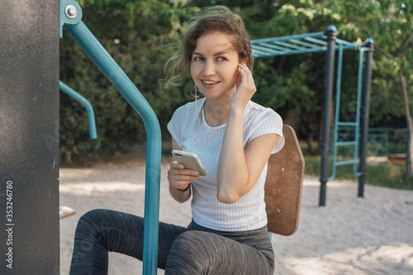 Fototapeta Girl brunette with blue eyes in a white top looks to the side holding a mobile phone and listens to music middle hand plan near the earpiece