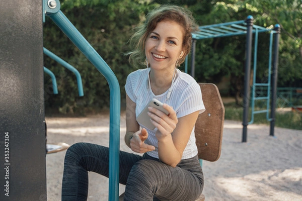 Fototapeta Girl brunette with blue eyes in a white top looks into the camera holding a mobile phone and listens to music medium plan