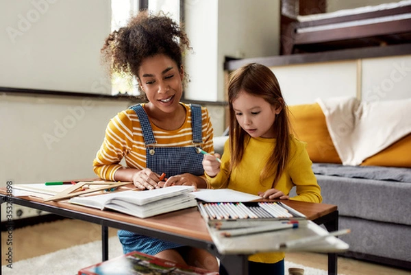 Obraz Responsibility and Reliability. Caucasian little girl spending time with african american baby sitter. They are drawing, learning to write letters, sitting on the floor