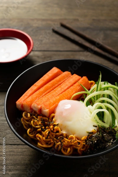 Fototapeta Spicy instant noodle with crab stick and onsen egg and cucumber in black bowl on wooden table.