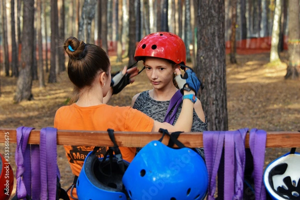 Obraz In a rope park, a beautiful young girl puts on equipment for traveling on a rope road, an instructor helps to put on a helmet, in a pine forest