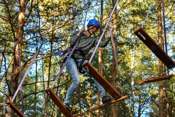 Obraz In a rope park, a beautiful young girl overcomes a difficult obstacle at high altitude, a shaky structure, a little scary and exciting, against the backdrop of huge trees and a clear sky, in a pine fo