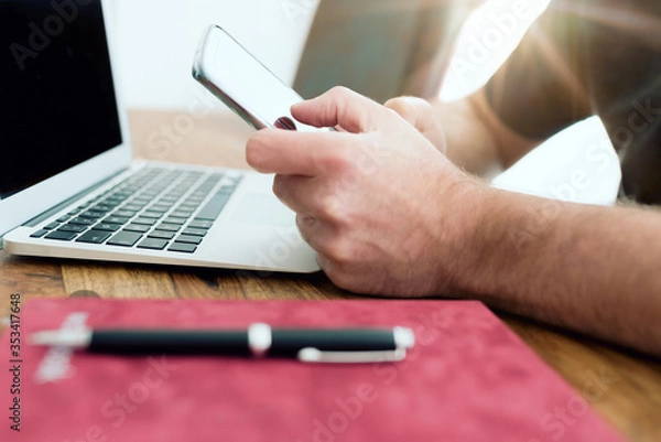 Fototapeta close-up of man using smartphone in front of laptop computer in home office