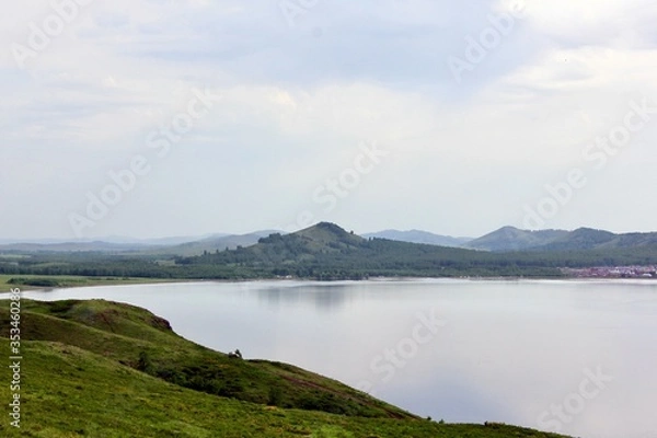 Fototapeta Panoramic view. A lagoon with a clean mountain lake in the midst of majestic mountains in a haze of fog. Green grass. Apartments around the lake