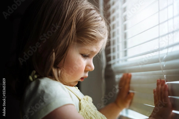 Fototapeta Little girl looking out  the window through the blinds. A child in quarantine at home.  
