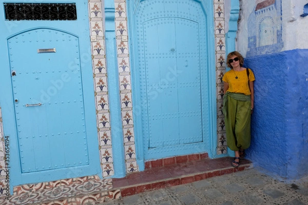 Fototapeta Pretty smiling woman standing near ornamented door in the blue city of Chefchaouen, Morocco.