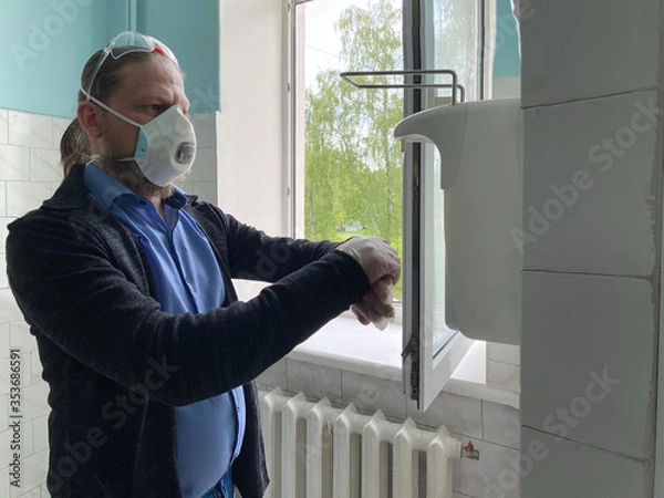 Fototapeta A man in a respirator and medical gloves treats his hands with an antiseptic. A man clicks on a dispenser for an antiseptic in a hospital after receiving a doctor.