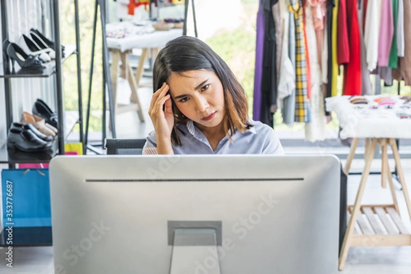Obraz Young Asian woman business owner sitting at a desk in a clothing shop looking at a computer monitor with a stressed worry unhappy headache expression on her face.