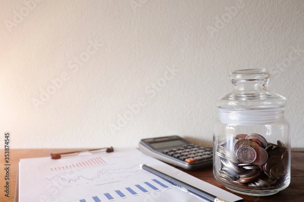 Fototapeta The cropped view of the glass jar contains the coins withe calculator and business paper on the wood desk with white natural blackground for texing the message, information,and background.