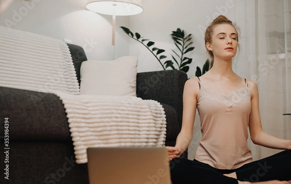 Fototapeta Red haired woman with freckles meditating on the floor using a laptop near the sofa