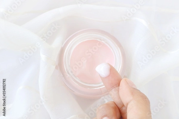 Fototapeta Top view of close up woman finger applying pink cream in jar on white fabric background