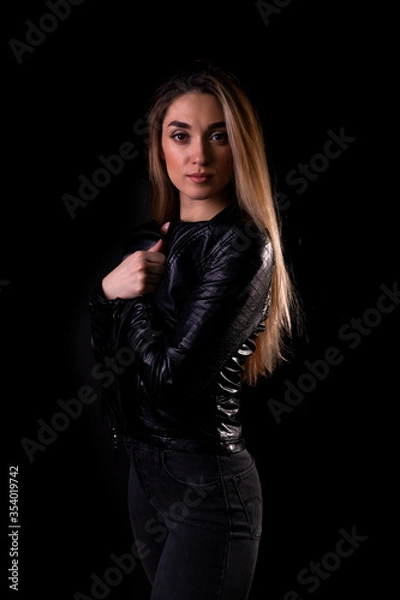 Fototapeta A young girl in a black leather jacket isolated on a black background