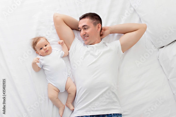 Obraz Happily family: father with his son a boy lie on their backs on a white bed in a sunny bedroom. Parent and little child are resting at home. View from above