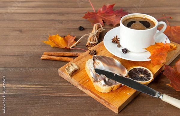 Fototapeta Cup of black coffee with a saucer, orange autumn maple leaves, cinnamon sticks, anise stars ,a piece of bread with creamy chocolate paste on a brown wooden background
