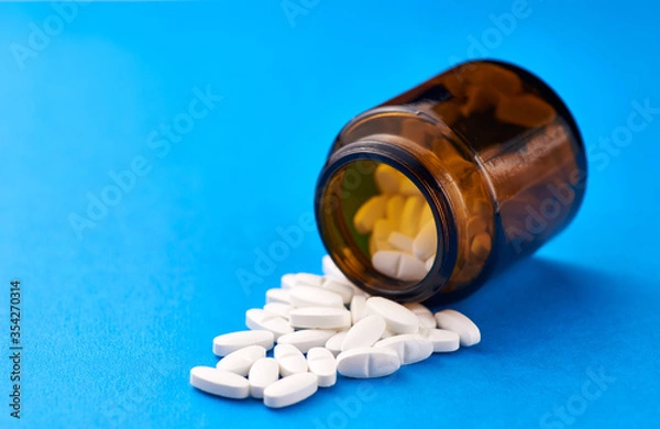 Fototapeta White pills from a brown bottle on a blue background.