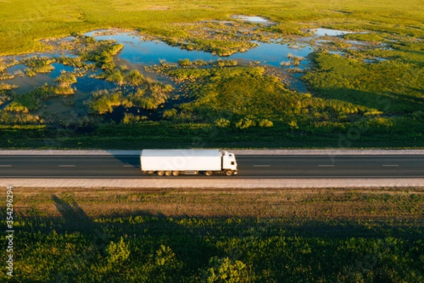 Fototapeta Semi truck with white freight trailer drive on highway in green grass countryside at sunrise, aerial top view. Car with motion blur effect. Road transportation and logistics background