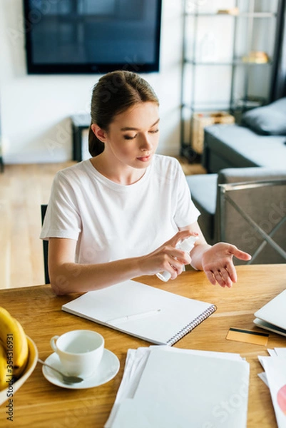 Fototapeta attractive woman holding bottle with hand sanitizer near notebook and cup on table