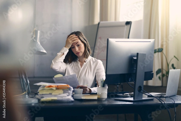 Fototapeta Very stressed business woman sitting in front of her computer looking at a large pile of paperwork, while holding a hand at her forehead 