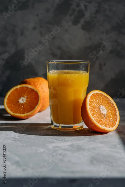 Fototapeta Orange juice in a glass, composition with halves of an orange with hard side sunlight on a gray background